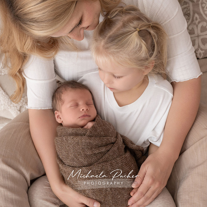 Mama sitzt mit ihrer kleinen Tochter auf dem Schoß, beide halten das schlafende Neugeborene, das in eine braune Decke eingewickelt ist.